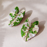 Two Alocasia Earrings shaped like monstera leaves with green and white enamel detailing are placed on a light surface, casting soft shadows—perfect for those who love lightweight jewelry.