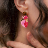 A close-up of a person’s ear wearing bold Anatomical Heart Earrings crafted from pink and gold holographic acrylic. Their curled brown hair partially frames the ear, while their hand gently touches their neck.