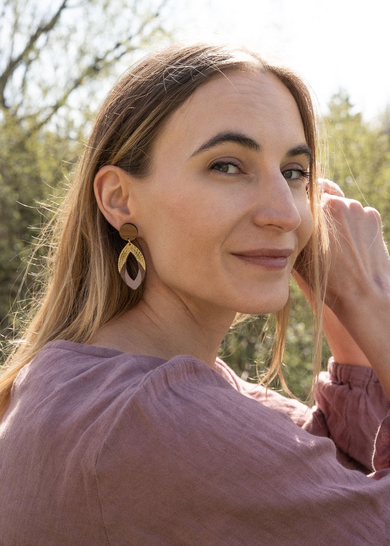 A woman with long light brown hair sits outdoors, smiling slightly as she looks over her shoulder. She wears a mauve blouse and handcrafted Bear Earrings made from premium acrylic glass, with sunlight and greenery in the background.