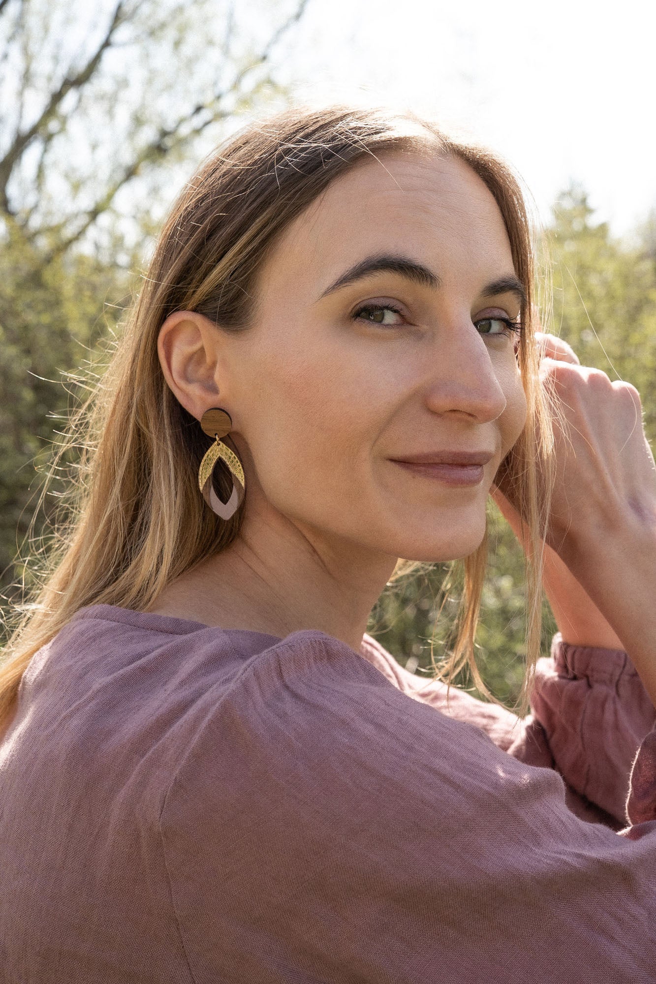 A woman with long light brown hair sits outdoors, smiling slightly as she looks over her shoulder. She wears a mauve blouse and handcrafted Bear Earrings made from premium acrylic glass, with sunlight and greenery in the background.