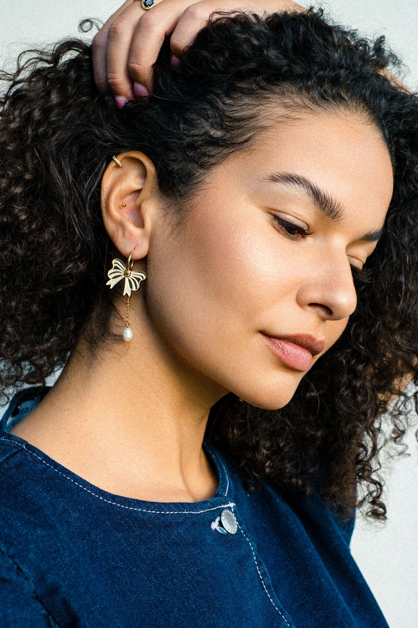 A woman with curly hair wearing a blue top looks down thoughtfully. She displays an interchangeable dangle earring with a dangling pearl on her left ear, her hand resting on her head.
