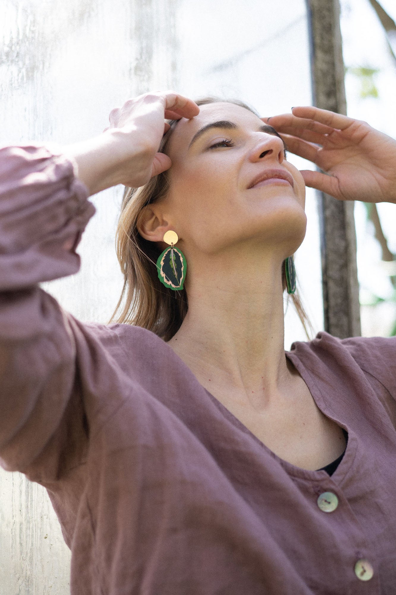 A woman in a mauve buttoned top stands outdoors by a glass wall, eyes closed and hands touching her forehead, wearing Calathea Medallion Earrings—nature-inspired jewelry that brings a relaxed elegance to her look in natural light.