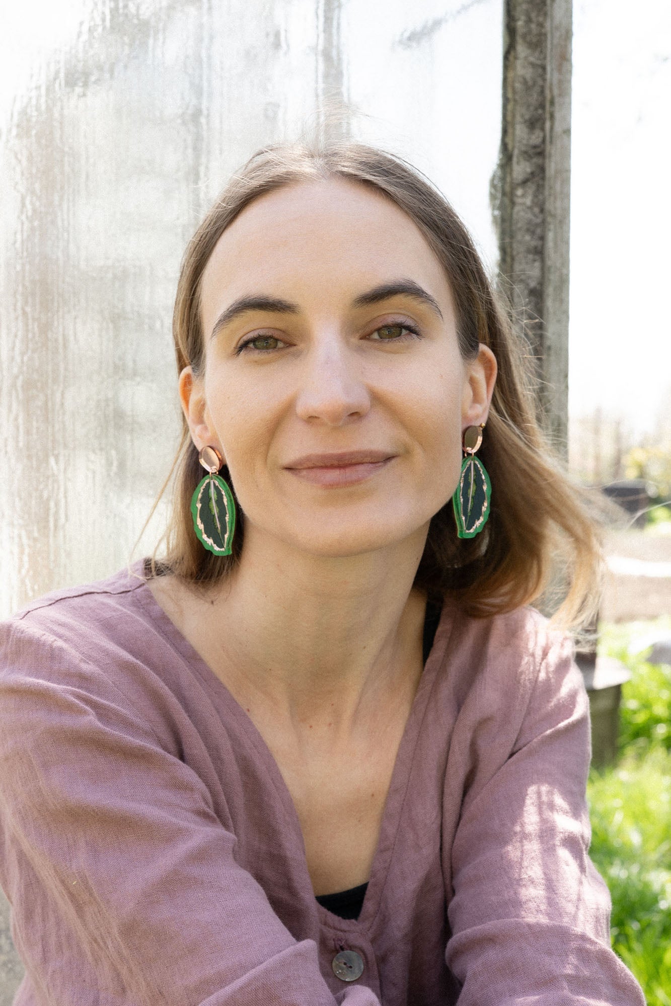 A woman with light brown hair, wearing a mauve blouse and Calathea Medallion Earrings, sits outdoors in sunlight, smiling softly at the camera. Blurred greenery and a glass panel create a serene backdrop for her nature-inspired jewelry.