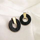 A pair of stylish Minimalist Circle Stud Earrings featuring black glittery acrylic glass hoops with gold rectangular accents, displayed on a white satin fabric background. Hypoallergenic for comfortable, all-day wear.