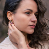 A woman with wavy brown hair wearing a beige top touches her Minimalist Circle Stud Earrings with one hand and rests her other hand on her collarbone. She looks slightly to the side with a calm expression. The earrings are hypoallergenic.