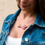 A woman wearing a denim jacket and white top looks down, showing a handcrafted gold necklace with a pendant shaped like a bird with outstretched wings and pink accents.