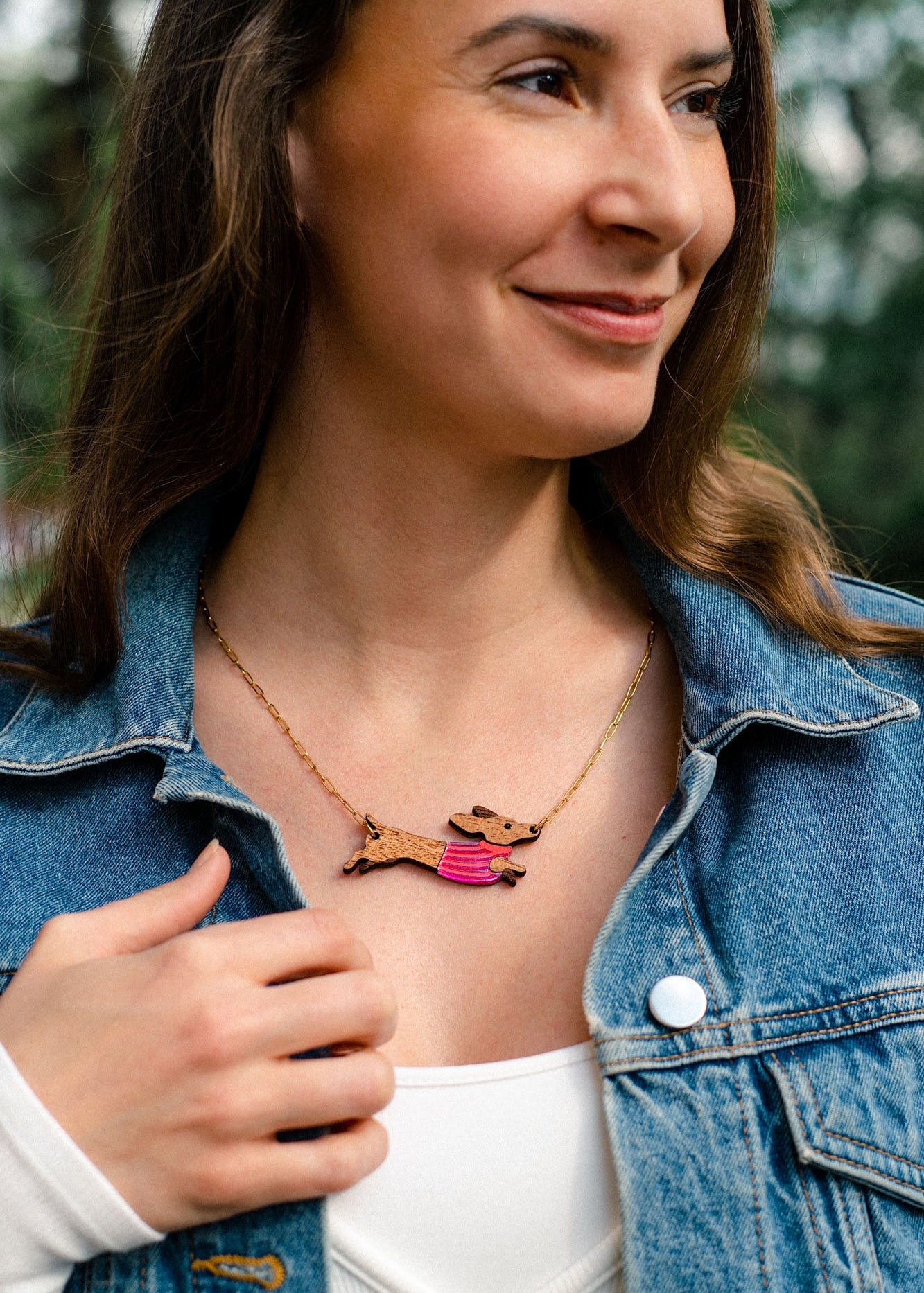 A woman wearing a denim jacket and white top smiles outdoors. Her long brown hair frames a colorful, handcrafted necklace featuring a Dachshund dog design made from sustainable wood.