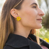 A stylish woman with long, straight hair reclines on grass outdoors, her unique accessories on display—Daffodil Stud Earrings adding charm as she looks peaceful, surrounded by blurred trees and greenery.