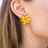 Close-up of a womans ear wearing Daffodil Stud Earrings with an orange center. Her light brown hair is tucked behind her ear, and she is dressed in a black top—perfect unique accessories for stylish women.