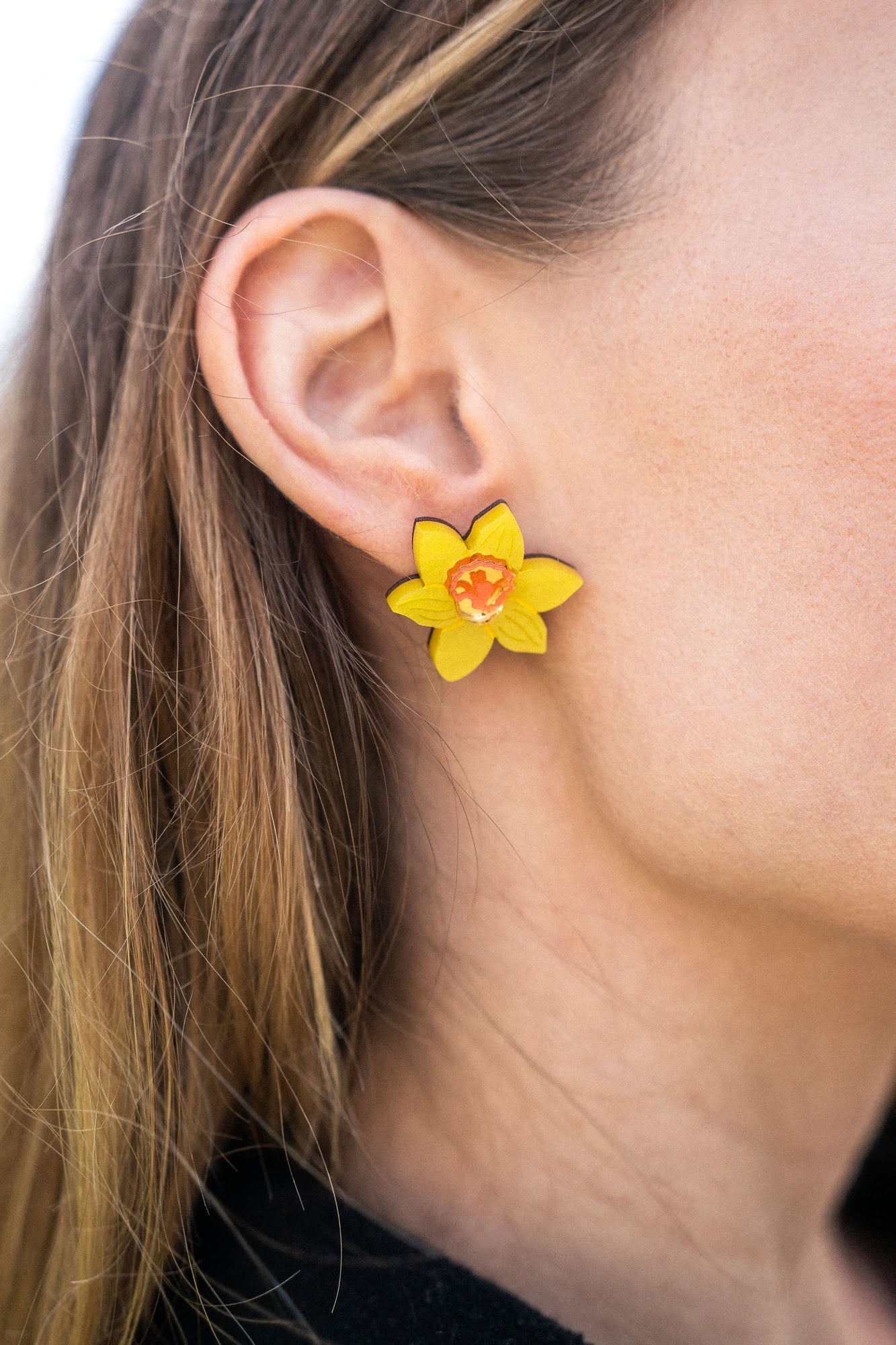 Close-up of a womans ear wearing Daffodil Stud Earrings with an orange center. Her light brown hair is tucked behind her ear, and she is dressed in a black top—perfect unique accessories for stylish women.