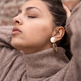 A woman wearing a beige turtleneck sweater and handcrafted gold earrings poses with her eyes closed, head tilted back, and hands resting behind her head in front of a brick wall.