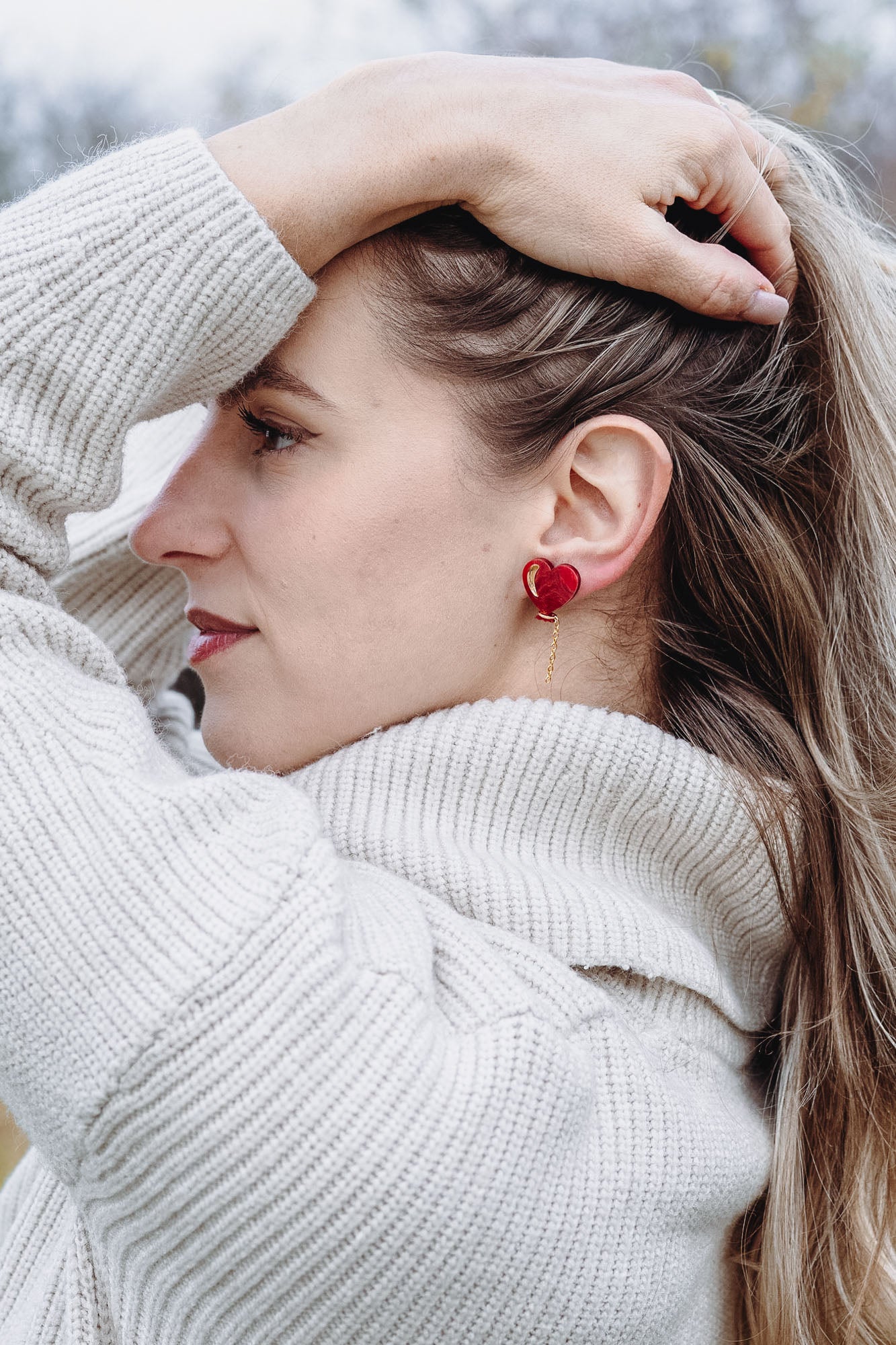 A woman in a cream-colored sweater stands outdoors, facing sideways with one hand holding back her long blonde hair. She wears playful Heart Balloon Studs, adding a romantic jewelry touch to her look. The background is blurry with trees and grass.