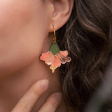 Close-up of a woman with wavy brown hair wearing handcrafted Hibiscus Earrings shaped like a pink and green flower, touching her neck gently with her fingers for a tropical statement look.