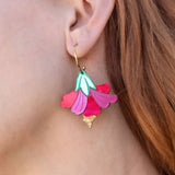 A close-up of a womans ear wearing handcrafted, petal-shaped Hibiscus Earrings in vibrant pink, teal, and gold tones. Her wavy, reddish-brown hair is partially visible, with tropical greenery softly blurred in the background.