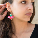 A woman with long brown hair wearing a black top and handcrafted, colorful Hibiscus Earrings looks to the side, touching her ear. The background is a light, textured wall.