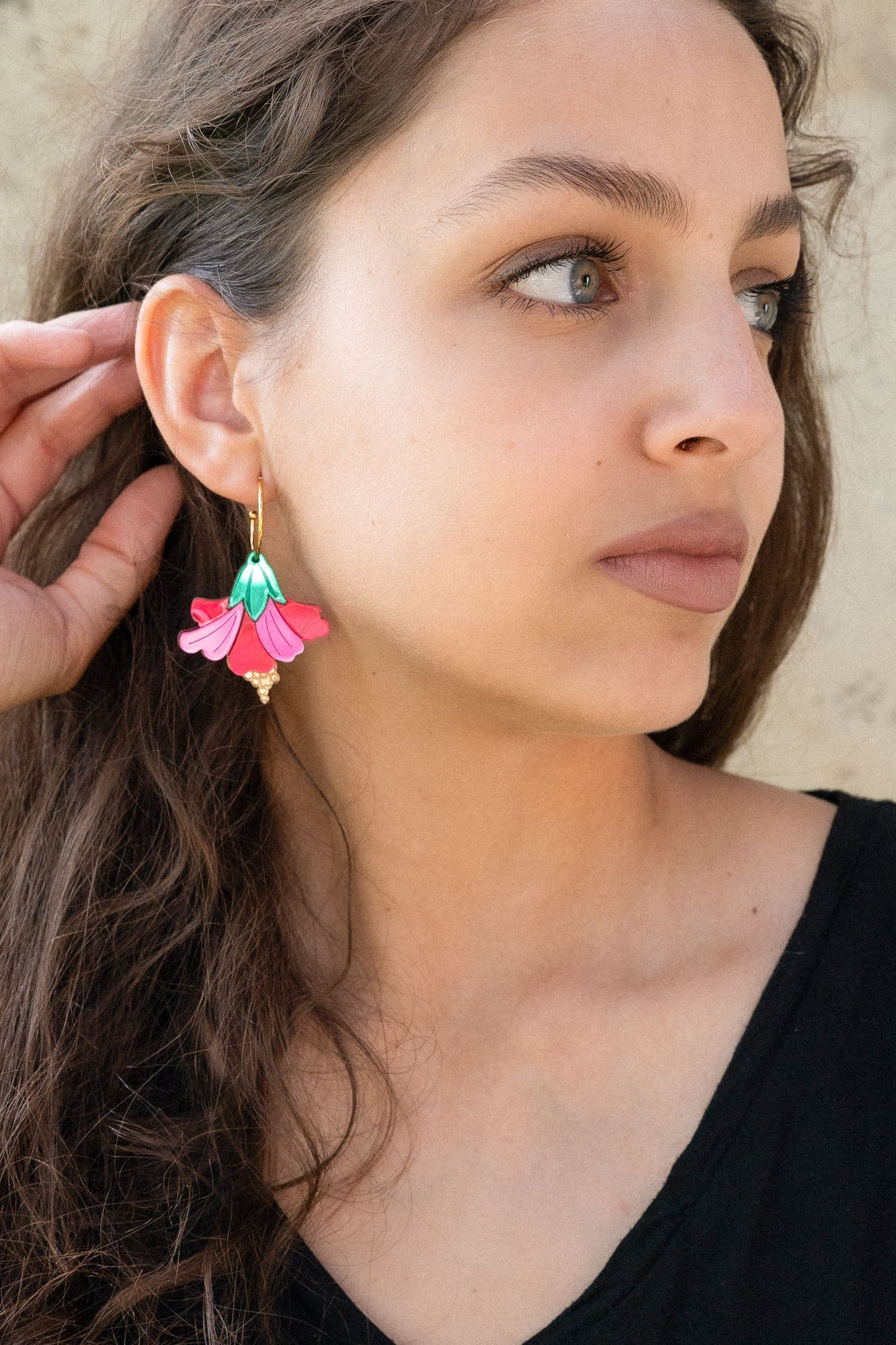 A woman with long brown hair wearing a black top and handcrafted, colorful Hibiscus Earrings looks to the side, touching her ear. The background is a light, textured wall.
