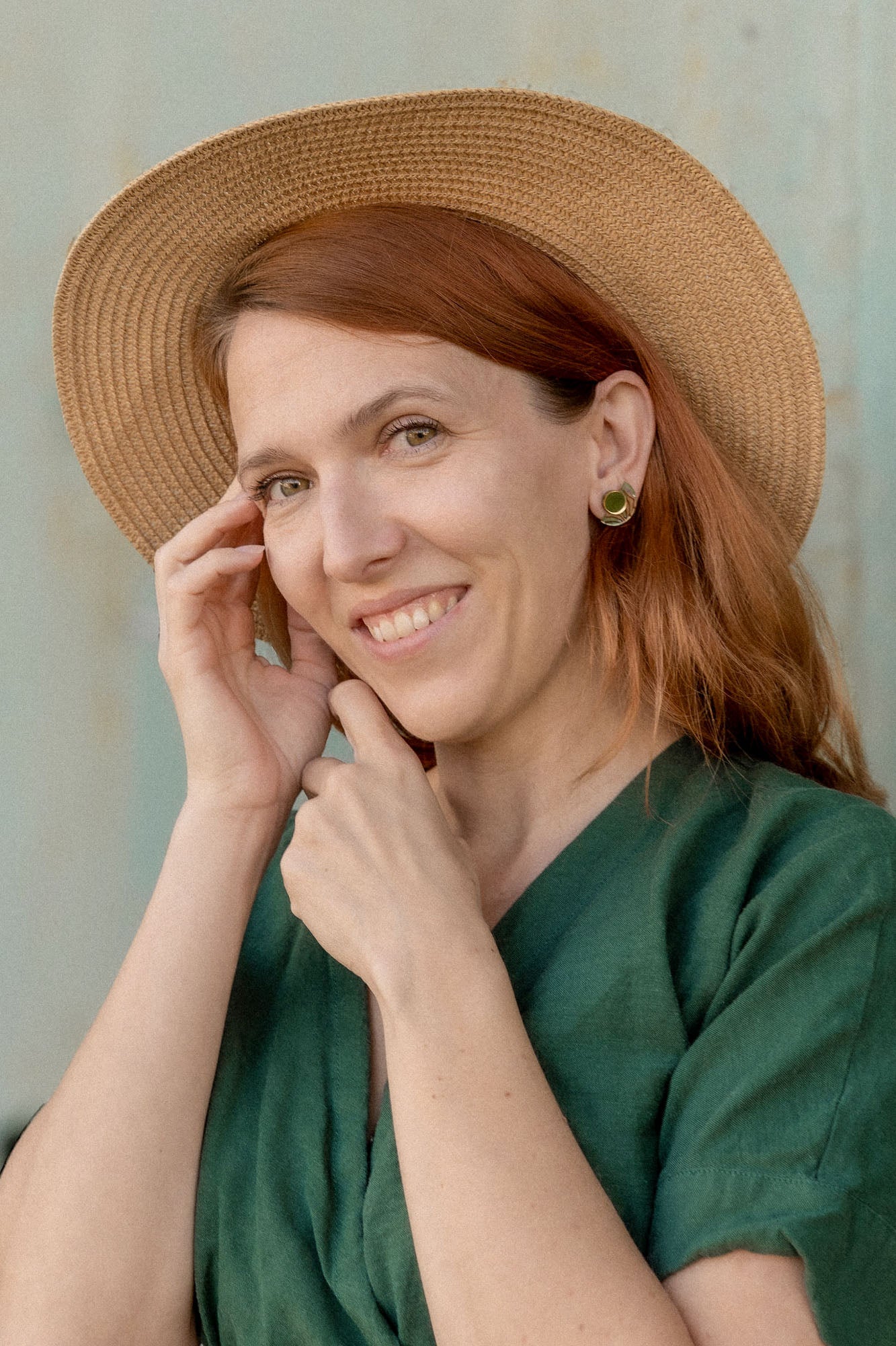 A woman with reddish-brown hair wears a green dress, a straw hat, and minimalist geometric stud earrings. She smiles gently at the camera while touching her face, standing in front of a light-colored background.