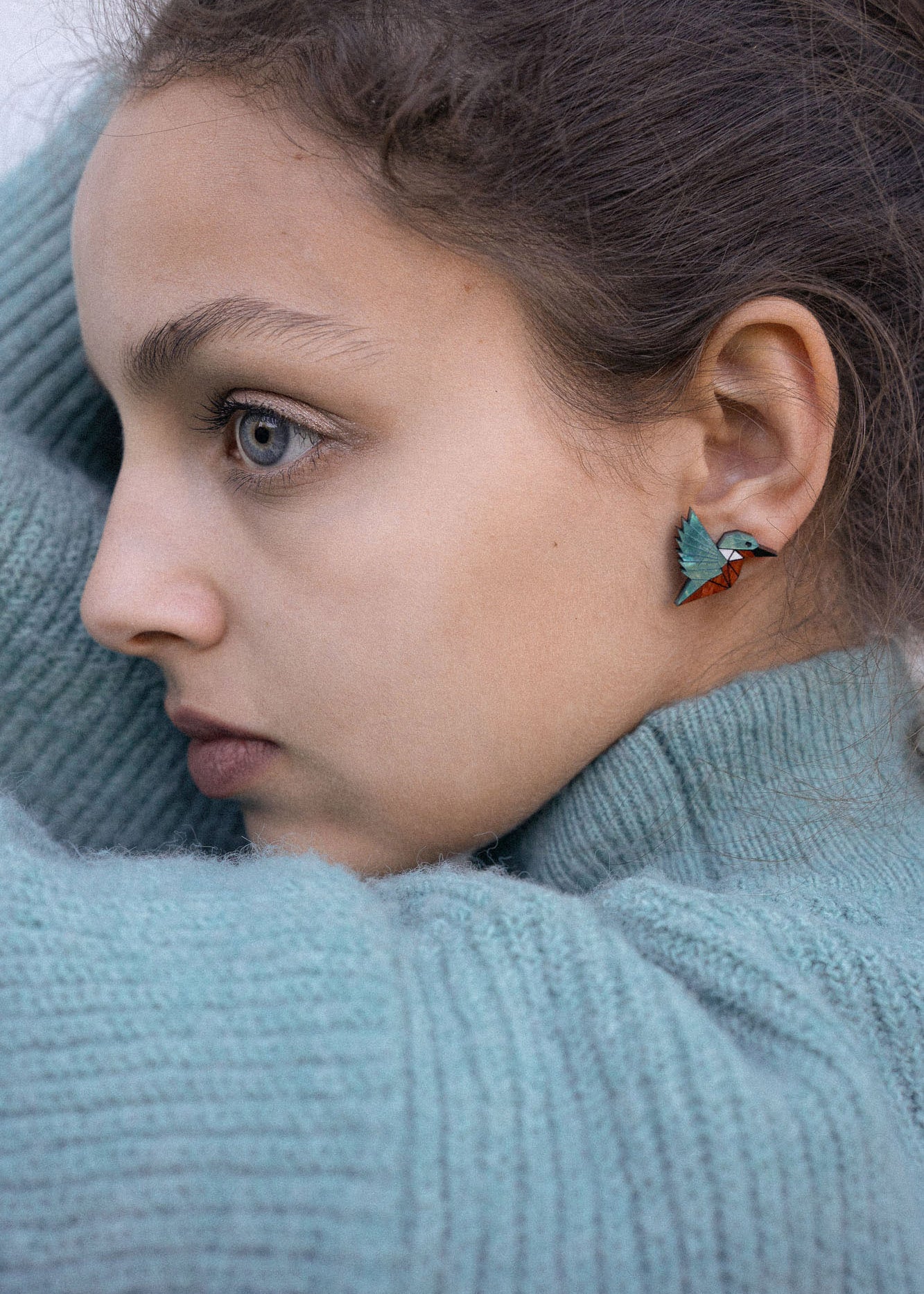 A young person wearing a teal knit sweater looks to the side, showing a Kingfisher Stud Earring. Their hair is pulled back, and their expression appears calm and thoughtful, embracing an eco-friendly, sustainably made accessory.