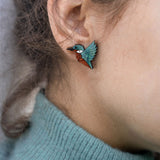 Close-up of a person wearing a turquoise sweater and eco-friendly Kingfisher Stud Earrings, shaped like a hummingbird, with the focus on their ear and earring. The background is softly blurred.
