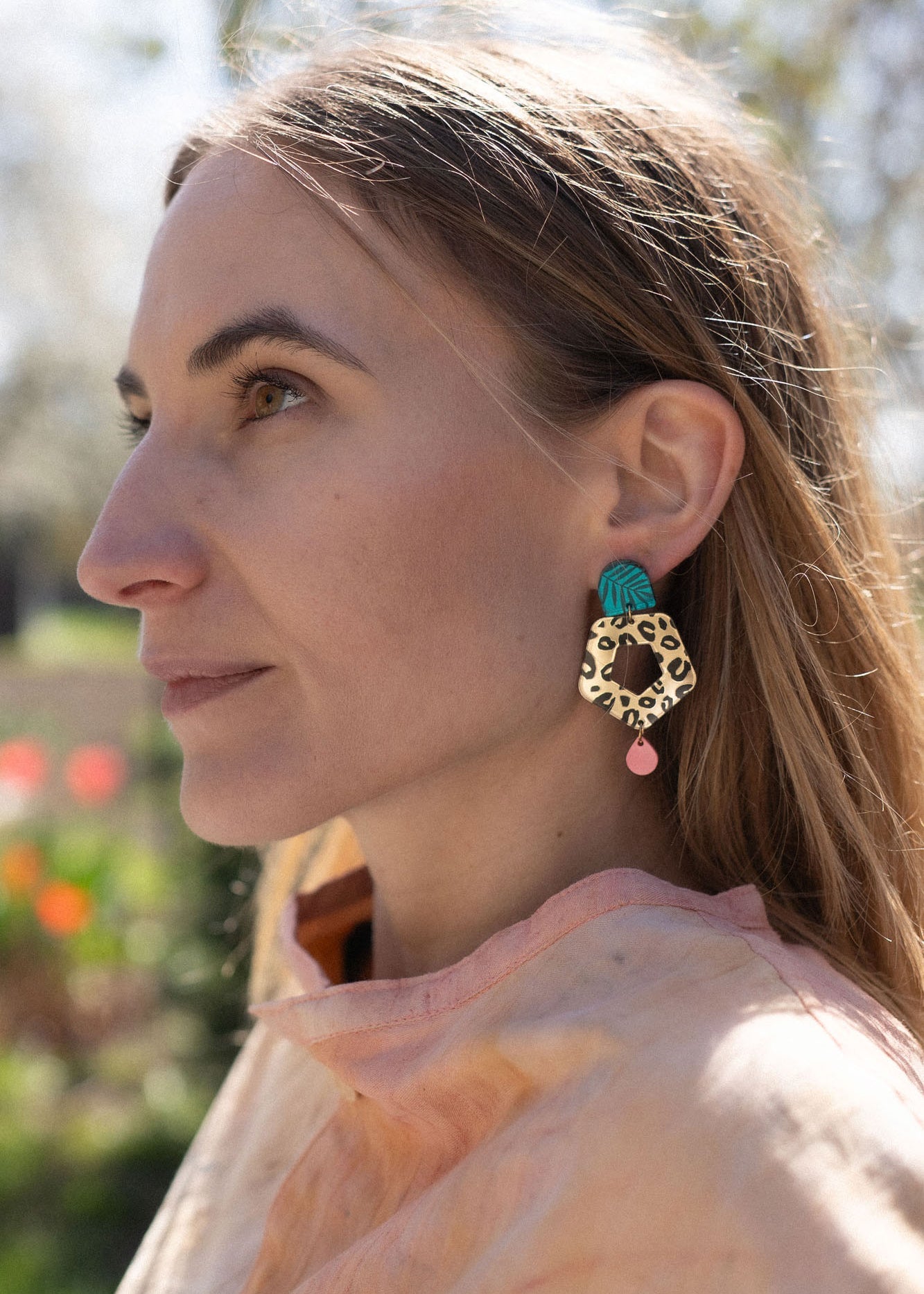 A woman with light brown hair gazes to the left, wearing a peach top and striking Leopard Earrings crafted from premium materials. The background is outdoors with blurred greenery and flowers.