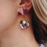 Close-up of a woman's ear wearing playful Mermaid Tail Earrings with a pink and purple heart-shaped stud and a semi-circular dangle in green, pink, and purple—a perfect accessory for mermaid lovers. The woman has blonde hair and wears a white top.