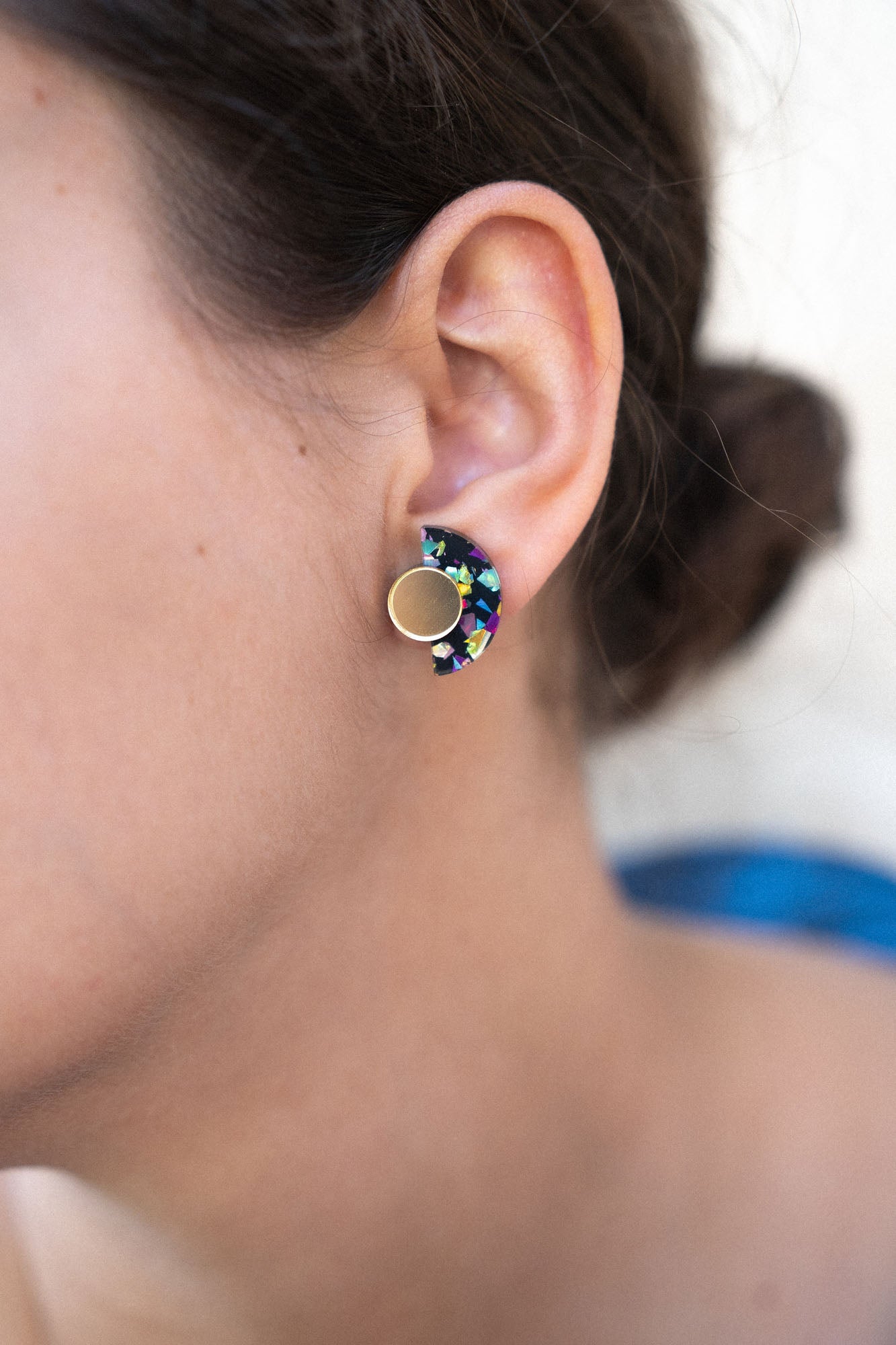 Close-up of a woman’s ear wearing colorful, crescent-shaped Minimalist Geometric Stud Earrings with a round gold stud. The womans dark hair is tied back in a bun. These hypoallergenic earrings are lightweight and set against a softly blurred background.