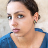A young woman with light eyes and dark hair pulled back, wearing a textured blue top and hypoallergenic Minimalist Geometric Stud Earrings, gazes seriously at the camera against a neutral, slightly textured background.