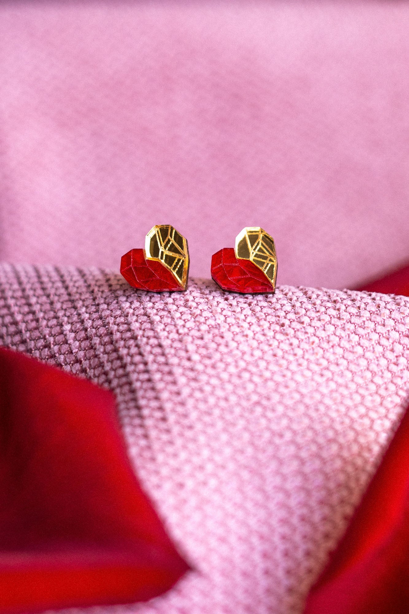 Gold and red geometric heart studs rest on pink and red textured fabric, with the colors and patterns creating a soft, romantic background—a perfect touch of romantic jewelry.