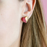 A close-up of a woman with light brown hair in a cream top, showing off romantic jewelry—a red and gold heart-shaped handcrafted earring. The pink background highlights the elegant design, with only the right side of her face visible.