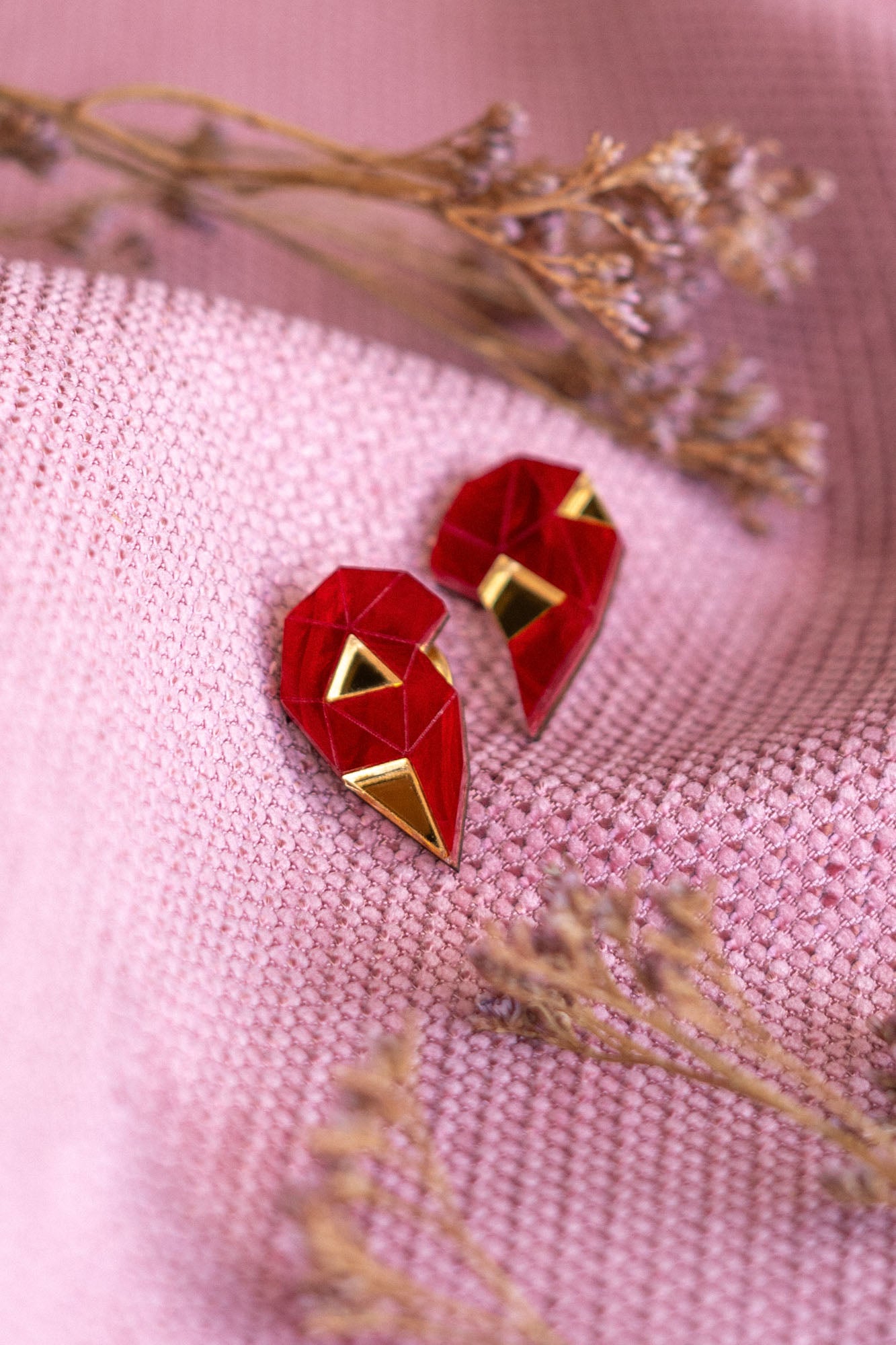 A pair of handcrafted geometric red and gold earrings shaped like hearts rest on textured pink fabric, with small dried flowers placed nearby—a perfect romantic gift.
