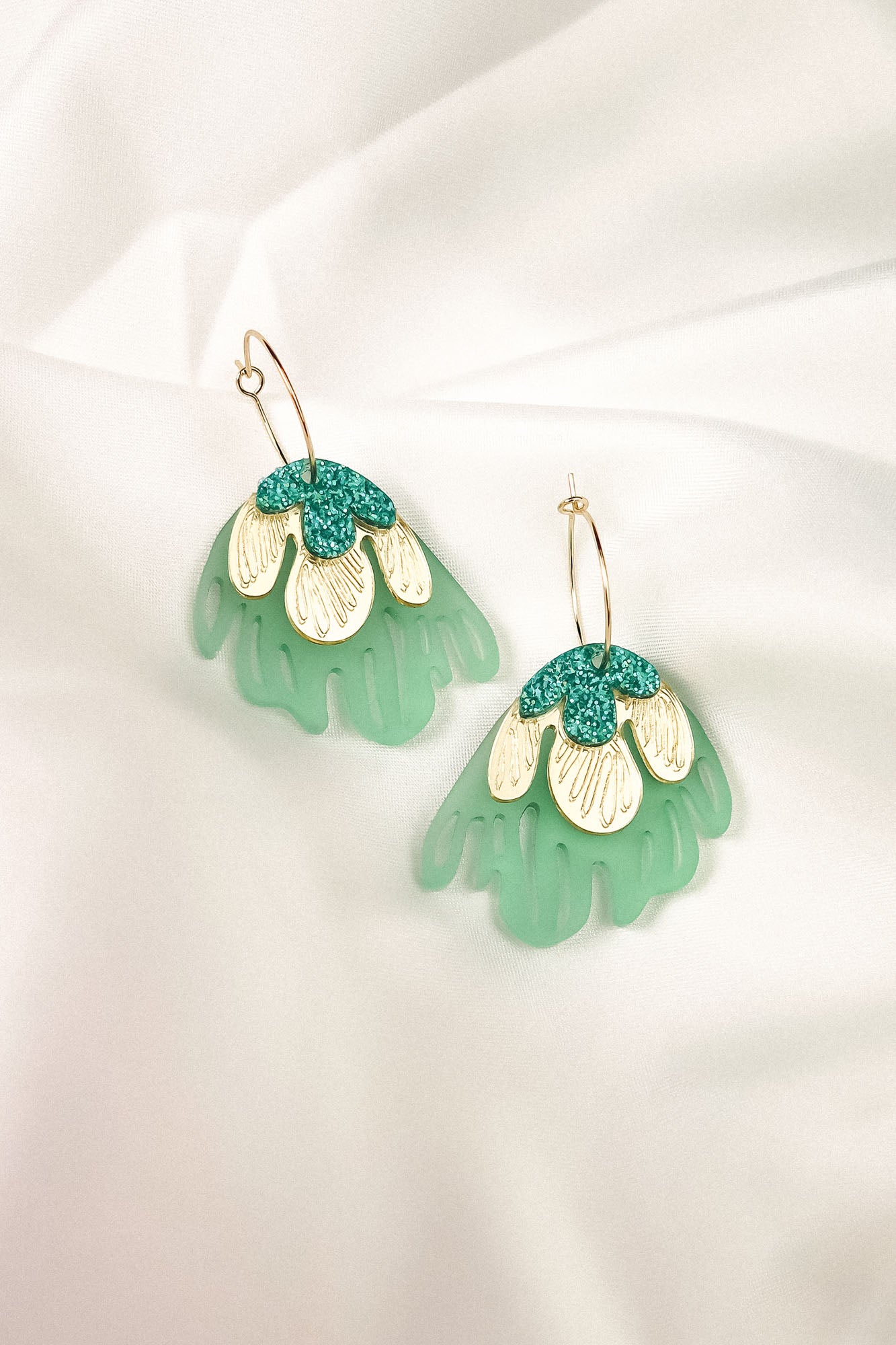A pair of handcrafted dangling earrings with gold hooks, featuring layered designs in mint green, cream, and teal glitter, resembling abstract floral shapes for a touch of tropical elegance, displayed on a white fabric background.