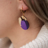 Close-up of a person wearing elegant purple plum earrings shaped like fruit with gold leaves. The person has light brown hair and is wearing a cream-colored, ribbed turtleneck sweater.
