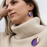 A woman with blonde hair wearing a cream-colored turtleneck sweater, handcrafted purple plum earrings, and a matching purple brooch stands outdoors, looking thoughtful with her hand resting on her head.