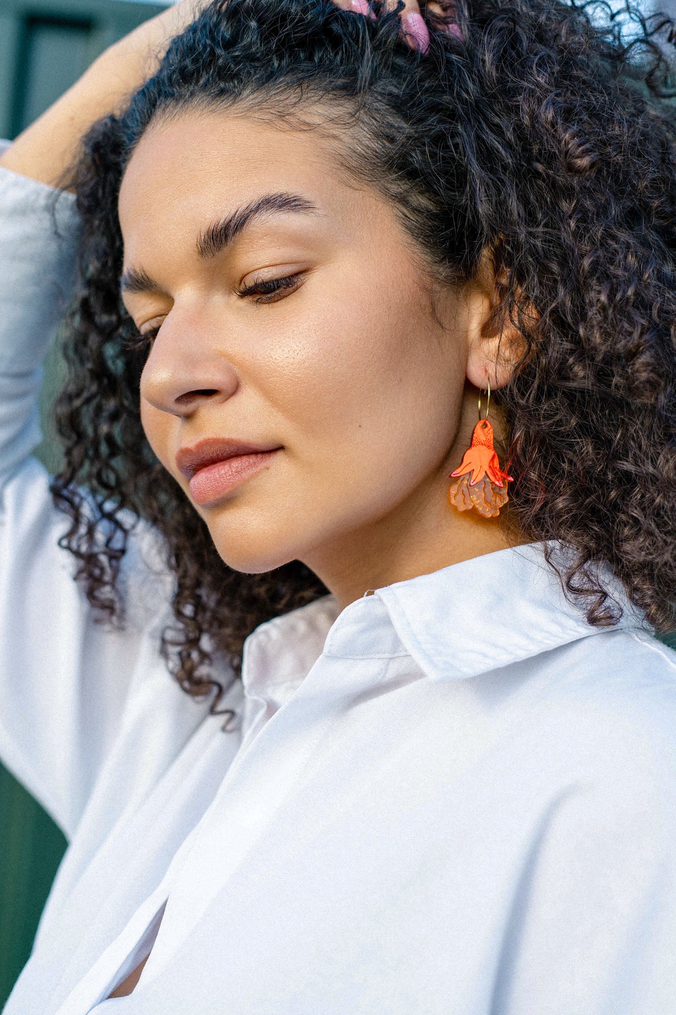 A woman with curly hair wearing a white collared shirt and handmade floral earrings looks down with a calm expression, her hand resting on her head.