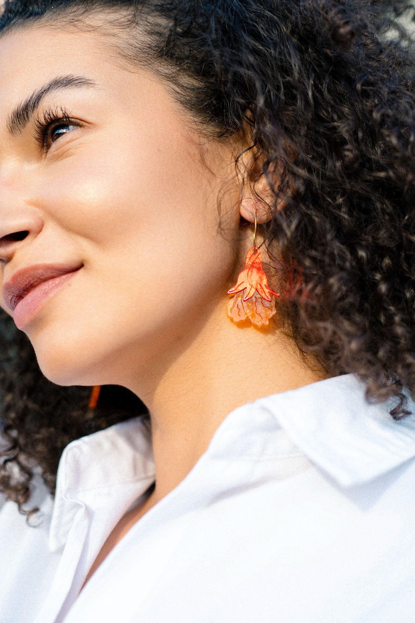 A woman with curly hair wearing a white shirt and handmade summer garden earrings smiles slightly while looking upward in natural light.