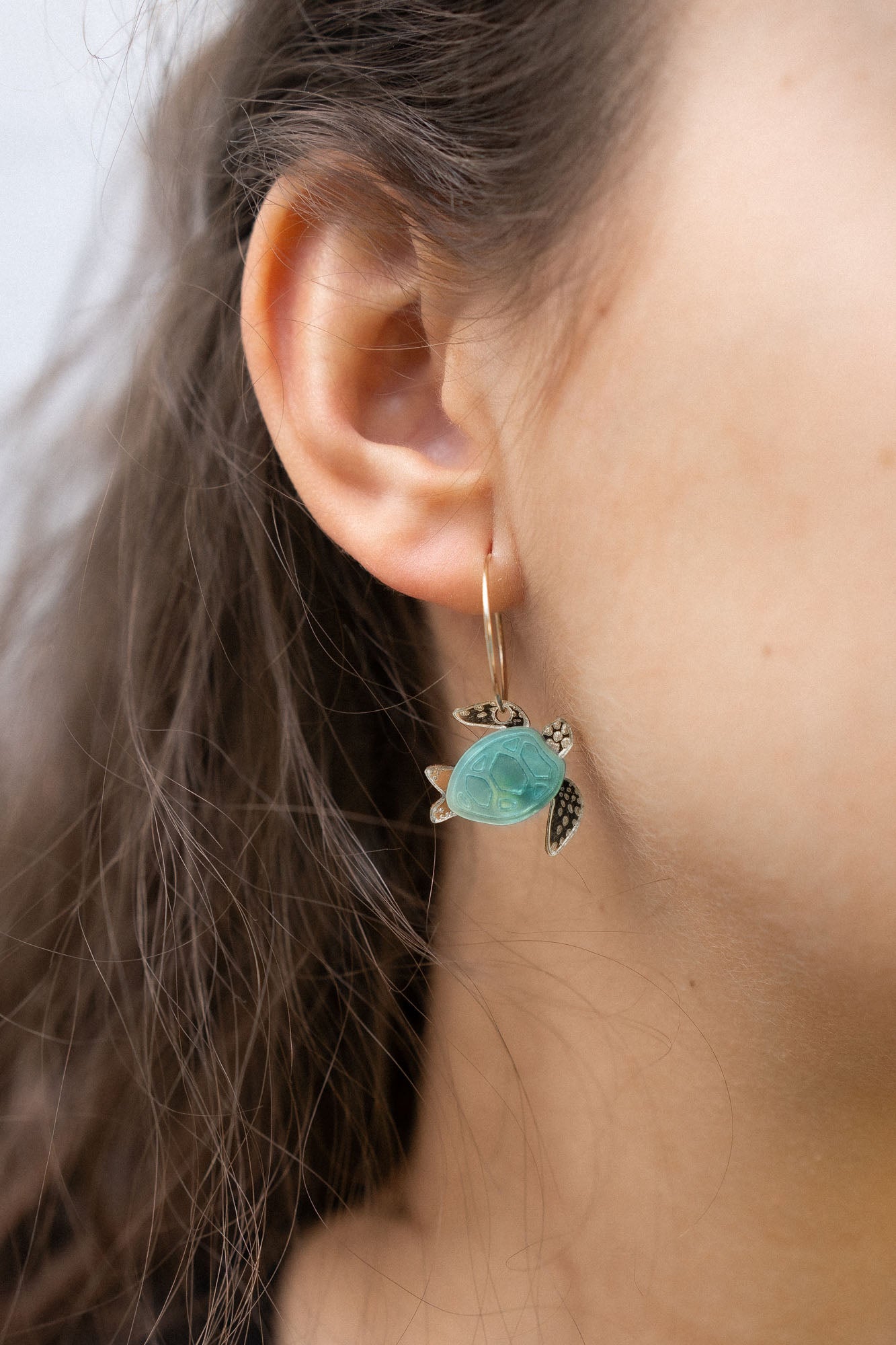 Close-up of a womans ear wearing Little Turtle Hoop Earrings—handcrafted gold hoops adorned with a turtle charm featuring a turquoise shell and gold flippers. The softly blurred background adds an elegant touch.