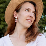 A woman with reddish-brown hair wearing a wide-brimmed straw hat, lightweight handmade seashell earrings, and a light V-neck top looks to the side, smiling softly. A blurred background of green trees is visible.