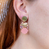 A close-up of a womans ear wearing a handmade gold stud earring with a bow and a dangling pink seashell-shaped charm. She has red hair, is wearing a straw hat, and the lightweight design adds delicate charm.