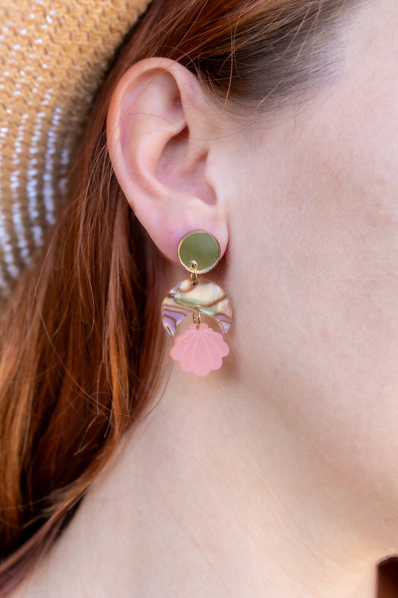 A close-up of a womans ear wearing a handmade gold stud earring with a bow and a dangling pink seashell-shaped charm. She has red hair, is wearing a straw hat, and the lightweight design adds delicate charm.