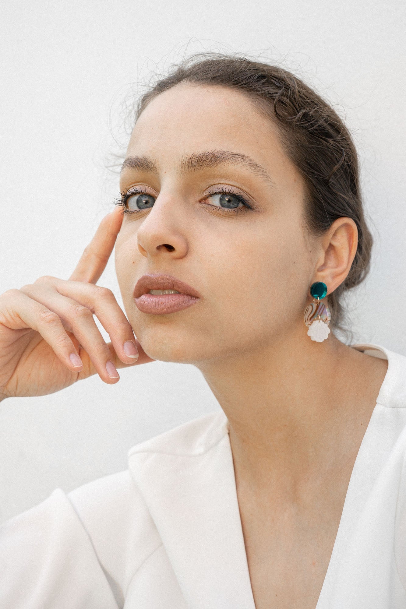 A woman with light skin and brown hair pulled back sits against a white background, wearing a white top and handmade seashell earrings. She rests her face on her hand, gazing thoughtfully at the camera.