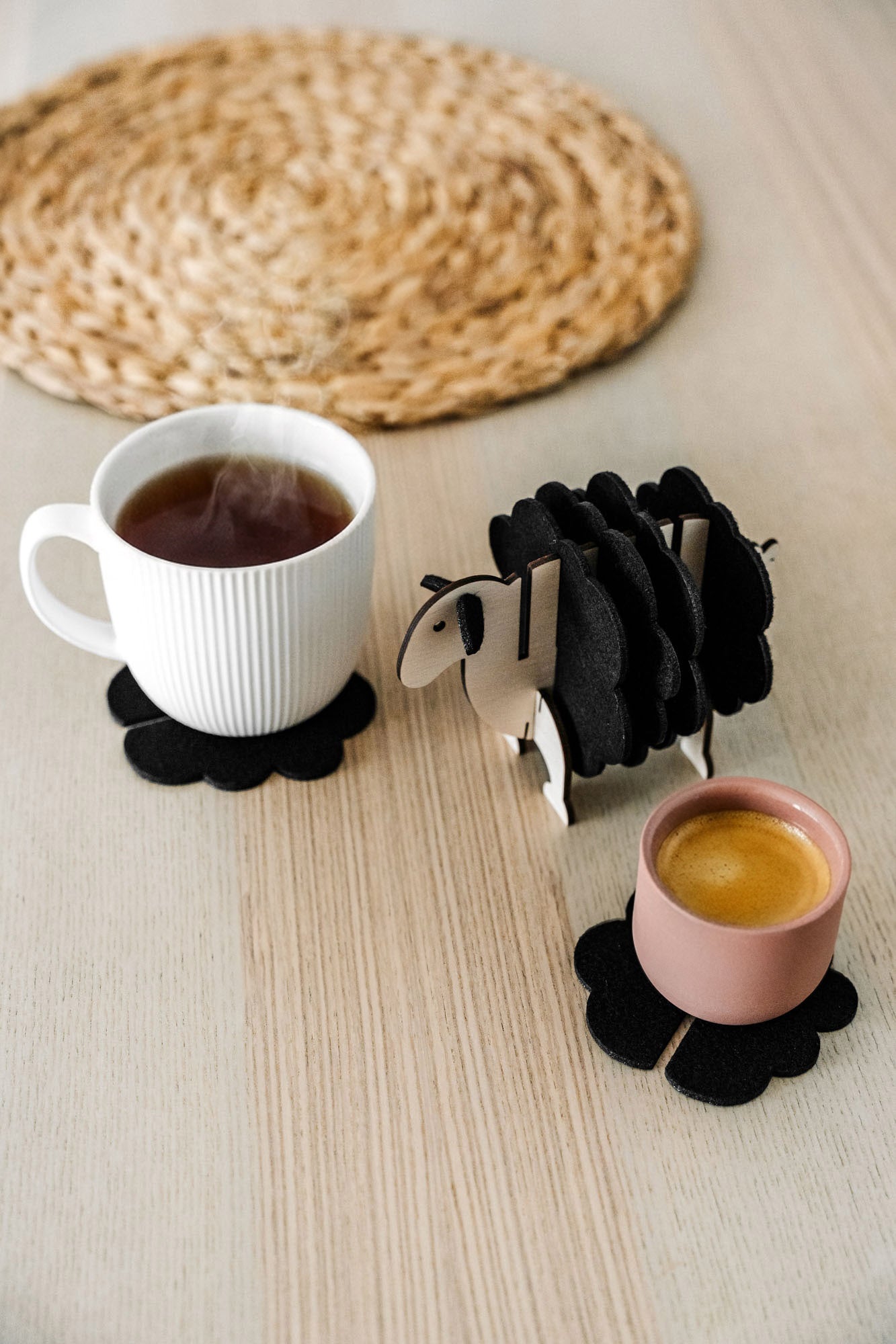 A white mug and a pink cup sit on black felt coasters next to a wooden sheep-shaped coaster holder on a light wooden table with a woven placemat in the background, perfect for charming home decor.