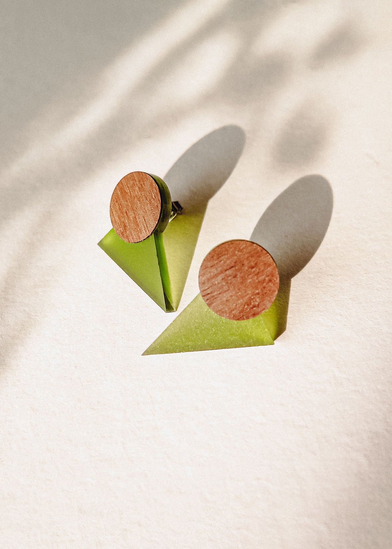 A pair of minimalist triangle earrings with round walnut wood tops and green acrylic glass bases are displayed on a light textured surface, casting soft shadows.