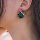 Close-up of a person’s ear wearing Tropicana Earrings—a tropical-inspired design with a gold tiger head, blue and pink details, and a delicate gold chain. The person’s hair is partially braided.