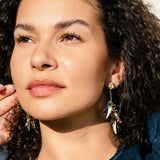A woman with curly hair and glowing skin gazes into the distance, wearing featherlight gold dangle earrings and a dark blue top. Sunlight highlights her features as she gently touches her hair with one hand.