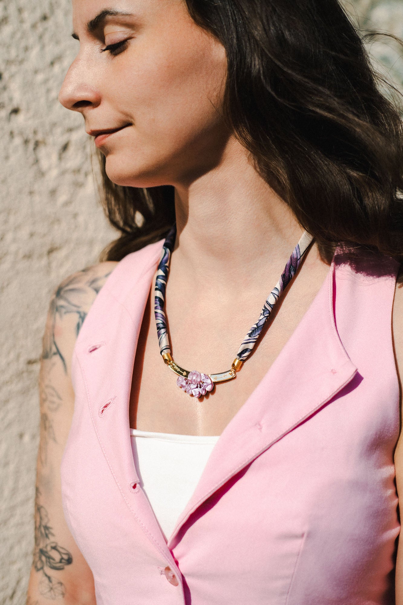 A woman with long dark hair and a floral tattoo on her arm wears a pink sleeveless vest, a white top, and a handmade necklace with a pink pendant, standing outdoors in sunlight.